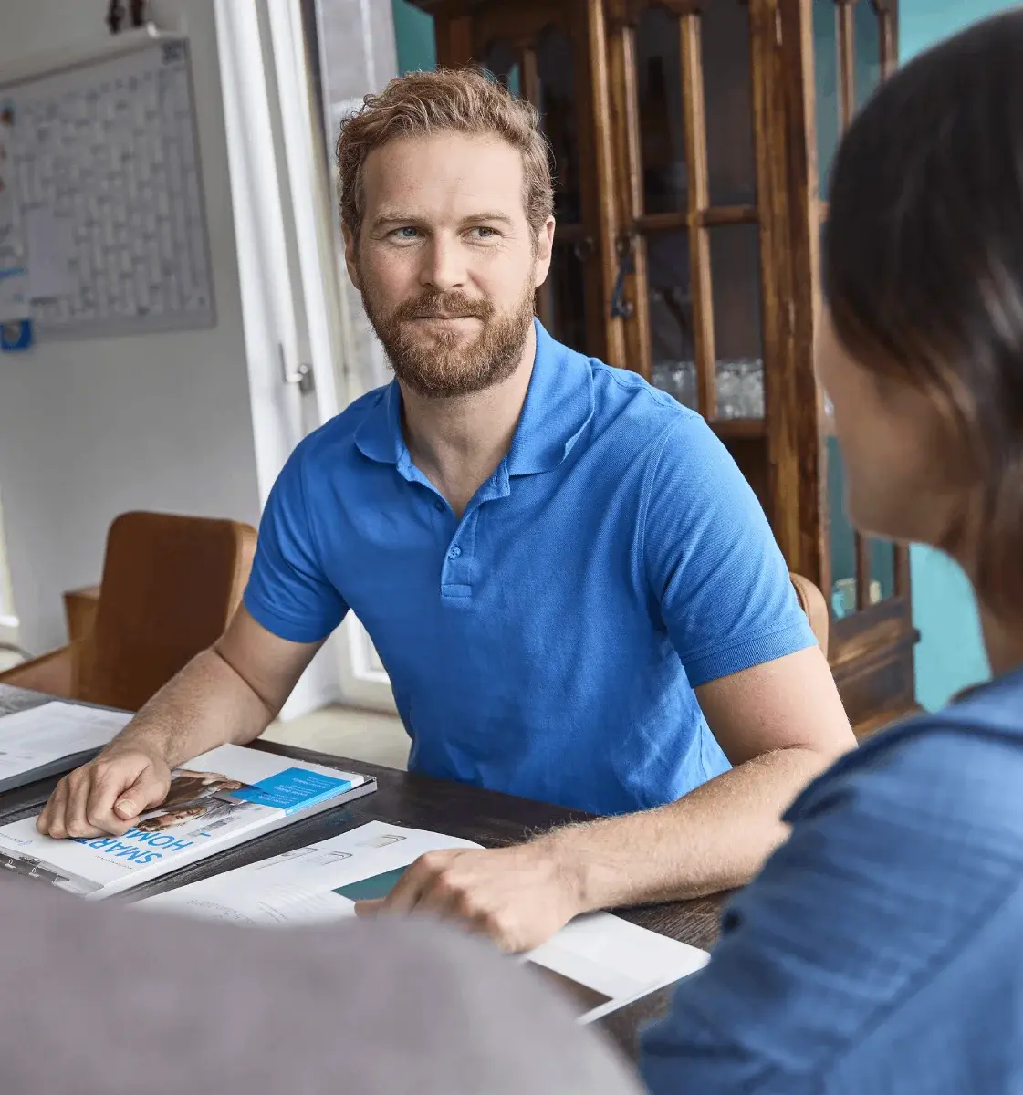 Ein älterer Mann in blauer Arbeitskleidung geht mit einem Ordner in der Hand an einem modernen Gebäude vorbei.