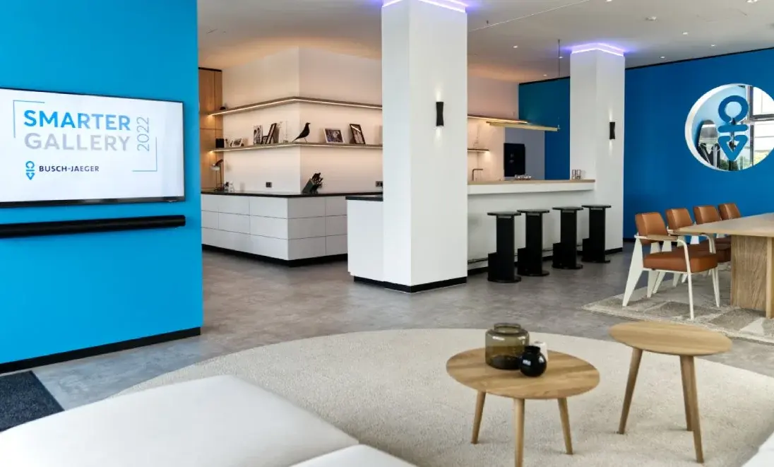 Moderne Innenansicht eines Raums mit blauen Wänden, einer Küche, Tischen und einem Bildschirm mit der Aufschrift „Smarter Gallery 2020“.