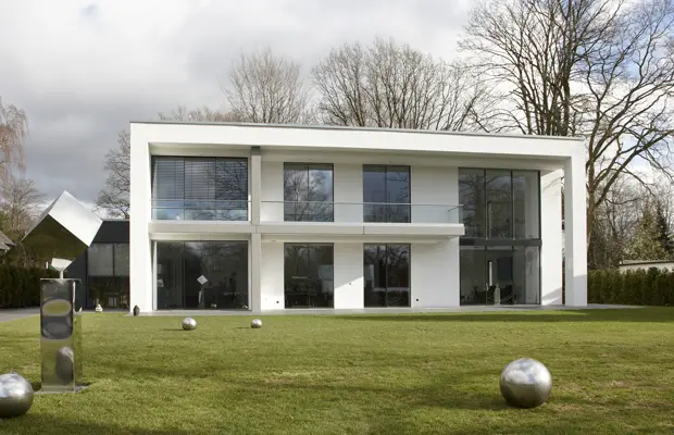 Moderne zweigeschossige weiße Villa mit großen Fenstern und einem gepflegten Garten, der mit runden Skulpturen geschmückt ist.