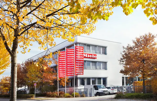 Miele-Gebäude mit roten Fahnen und autumnlichen Bäumen im Vordergrund.