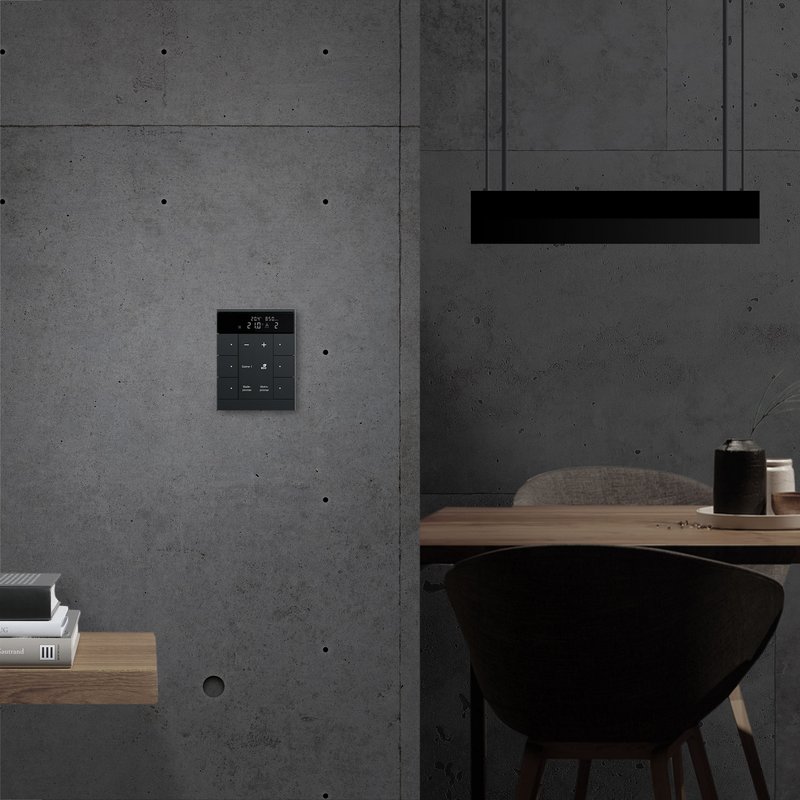 Intelligente Steuerungseinheit an einer Wand aus Beton, neben einem Tisch mit einem Stuhl und gestapelten Büchern.