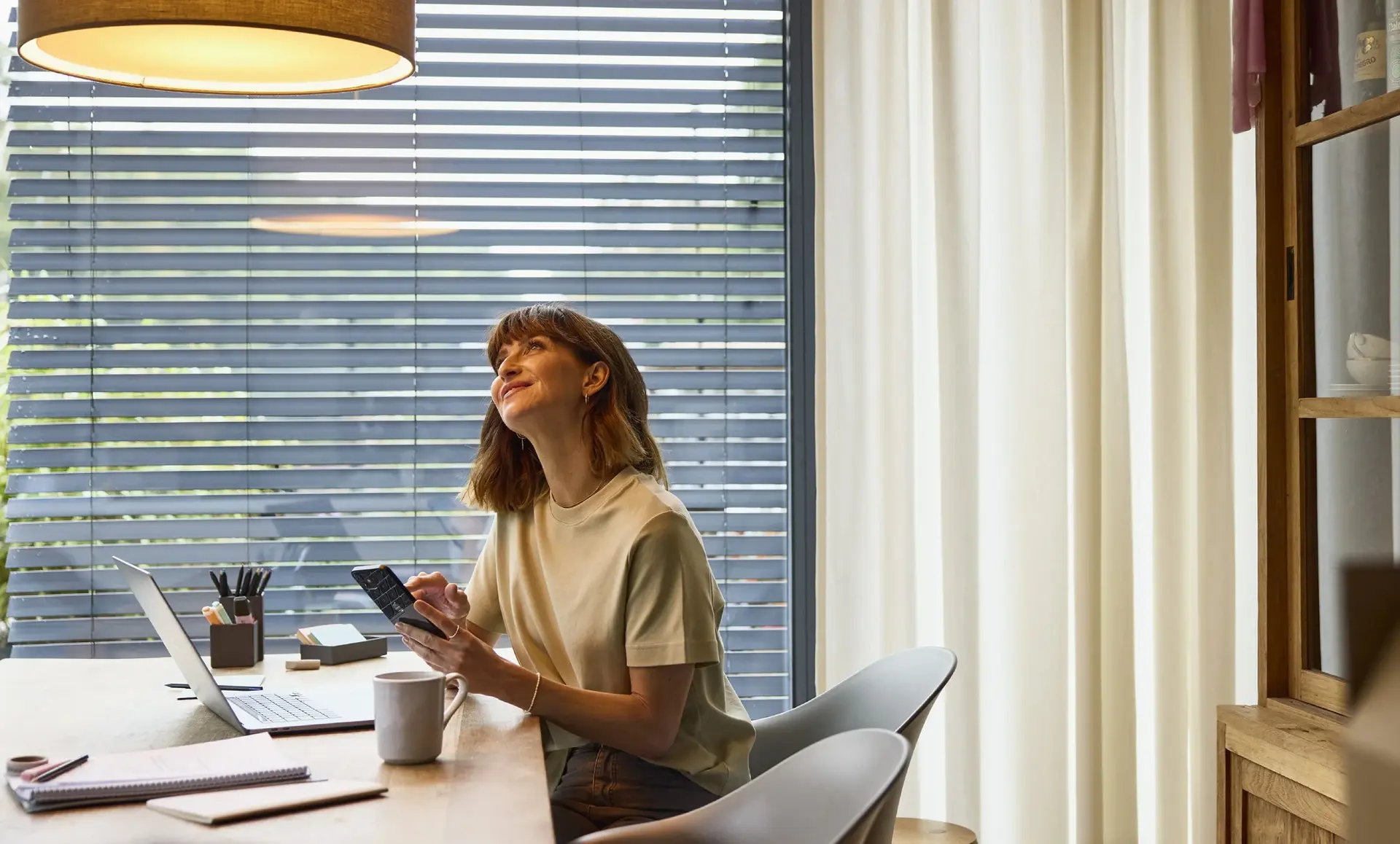 Frau sitzt an einem Tisch mit Laptop und Tablet, lächelt und blickt zur Lampe über ihr.