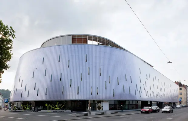 Moderner, gewellter Gebäudekomplex mit metallischer Fassade und großen Fensteröffnungen an der Straßenecke.