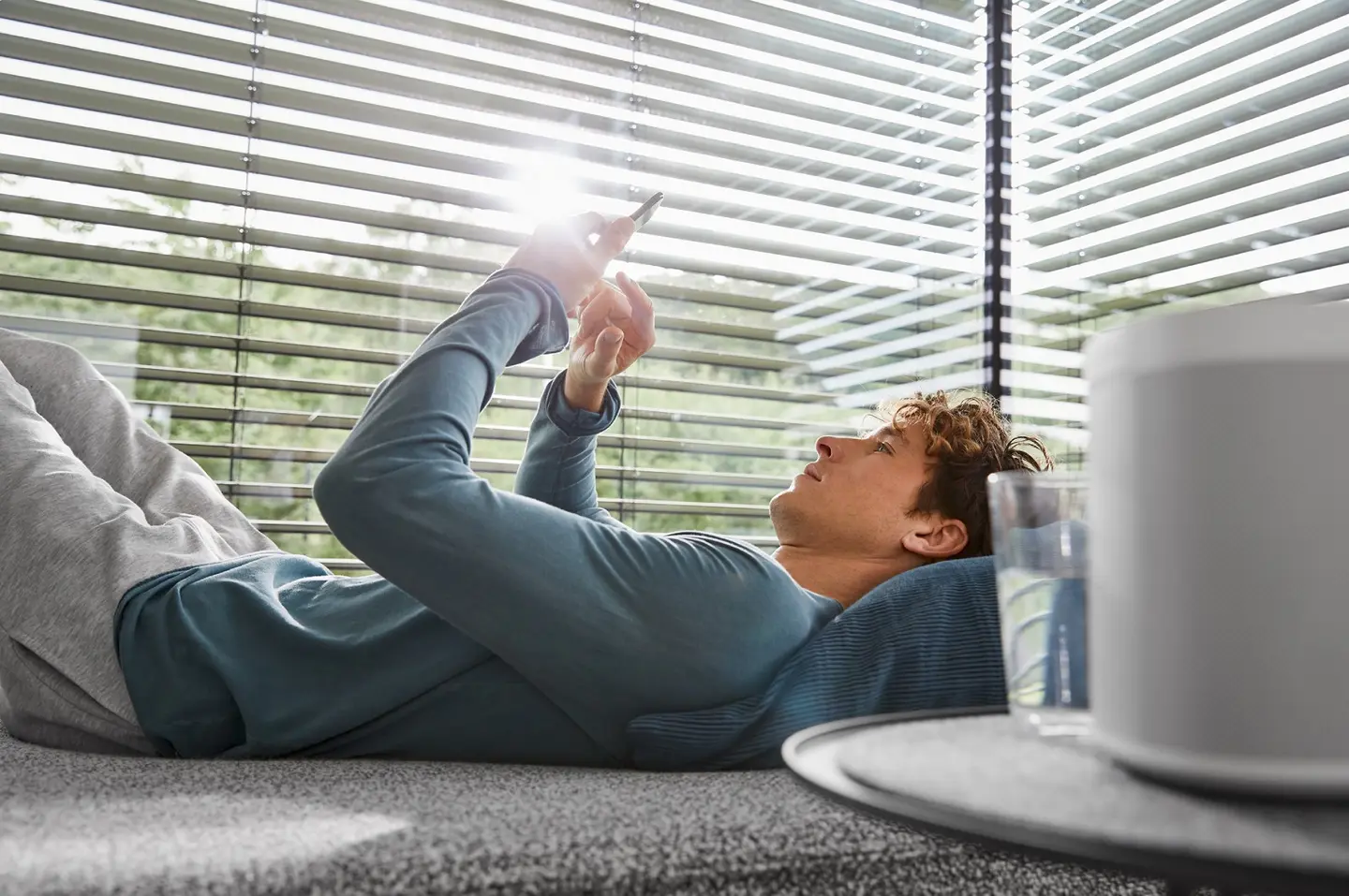 Junger Mann liegt auf einem Sofa und nutzt ein Smartphone, während Sonnenlicht durch die Jalousien strahlt.