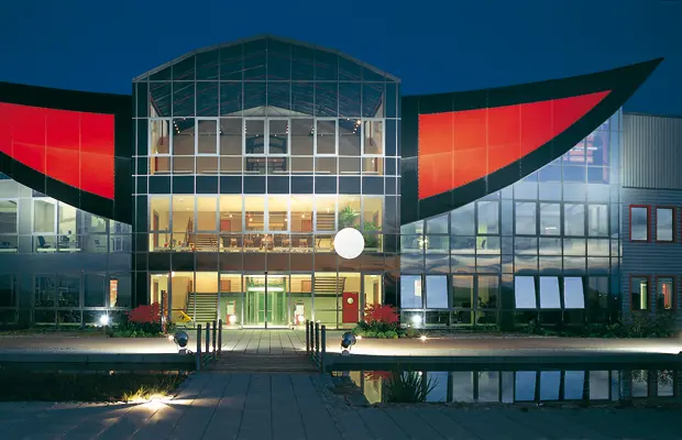Moderne Gebäude mit großflächiger Glasfassade und auffälligen roten Elementen, beleuchtet bei Nacht.